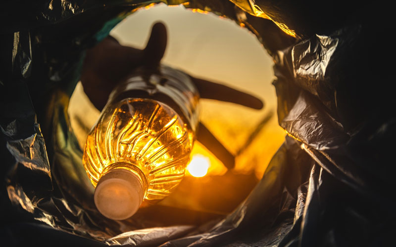 Photo of an empty plastic bottle being tossed into a plastic bag. The image is taken from inside the plastic bag, and one can see the outline of a hand tossing the bottle. Behind, a bright sunset shines through the litter.