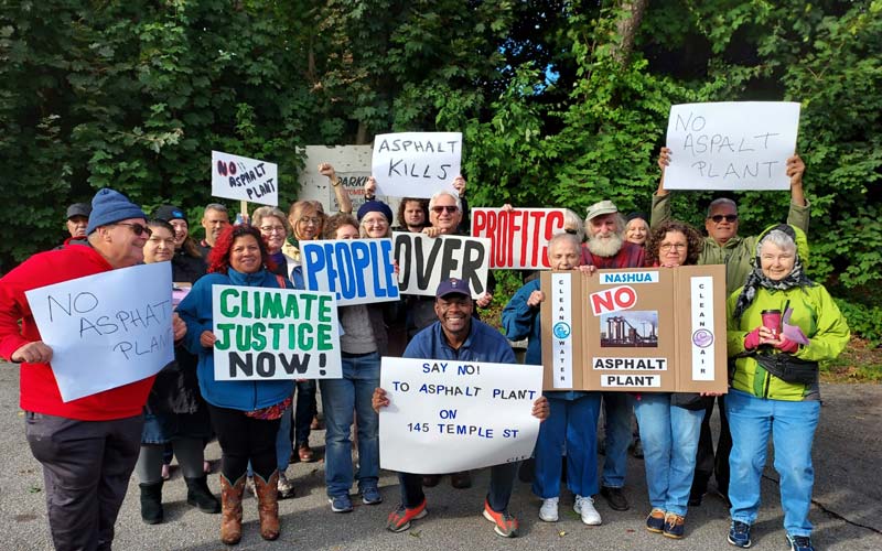 Photo: A group of diverse people is gathered outdoors, holding signs in protest against an asphalt plant. The signs display messages such as "No Asphalt Plant," "Climate Justice Now!" "Asphalt Kills," "People Over Profits," and "Clean Water, Clean Air." The protesters are standing together in solidarity, with trees providing a natural backdrop. The mood is determined and peaceful as the group advocates for environmental justice and public health. Proposed plant would be located in Nashua.
