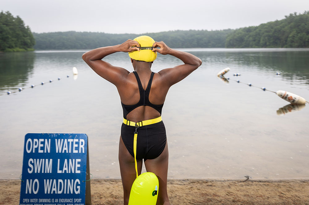 A Black woman in a black bathing suit and yellow swim cap gets ready to swim at a pond surrounded by green trees.