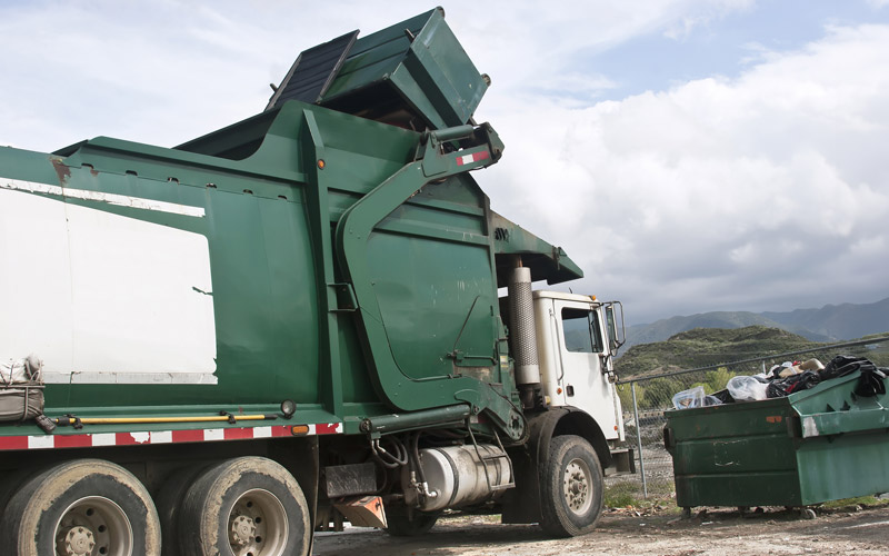 Photo: image of garbage truck carrying waste from a dumpster into a waste transfer station or a landfill.