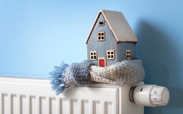 A small toy house wrapped in a scarf sits on a radiator against a blue wall. The house has a gray roof with four windows and a small red door.