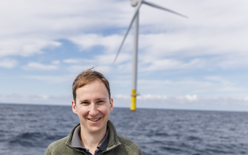 CLF's Nick Krakoff in front of one of South Fork's wind turbines. The sky is bright blue and cloudy. Nick is wearing an army green pull over sweater and smiling at the camera.