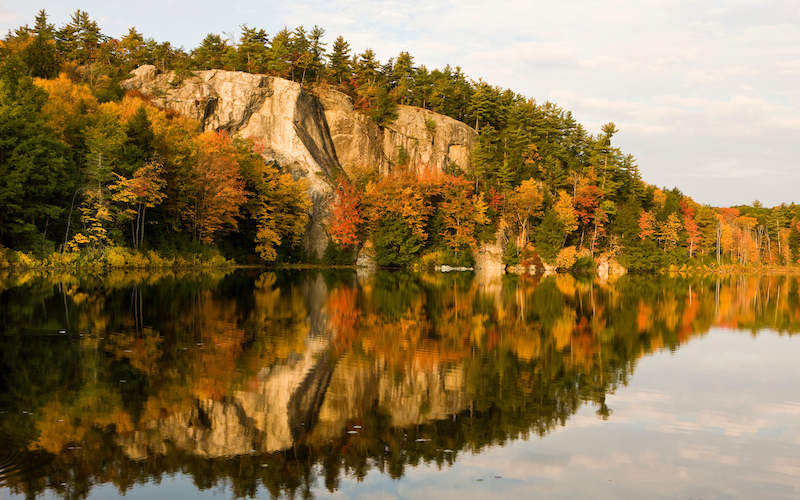 An angled view of a rocky mountain with trees covering it that are changing to their fall colors. Below the mountain is a body of water reflecting the scene above.