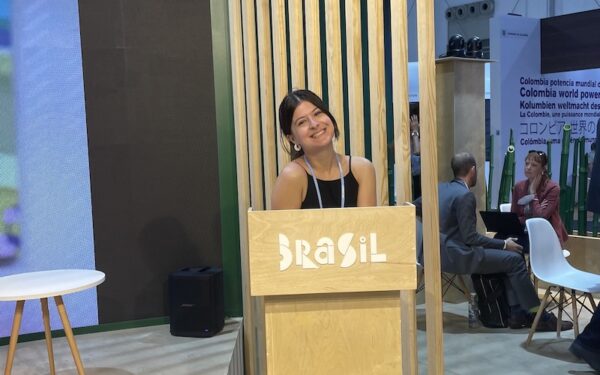 Thalia Chaves stands behind a podium with "Brasil" written on it in white lettering. She is wearing a black shirt and a lanyard and smiling at the camera.