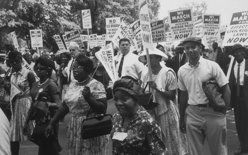 Civil rights protesters carrying sing. Photo is in black and white. Two black women wearing floral dresses stand at the frot. Protest called for higher minimum wage, stopping the jim crow era, and more. The march resulted in the creation of many of the provisions in Title VI of Civil Rights Act.