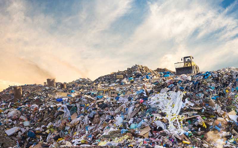 A sprawling landfill under a bright sky, with piles of mixed waste. A bulldozer sits atop the waste, processing the layers. The scene conveys the scale of waste disposal and the potential for strong smells often associated with landfill operations and the mix of garbage.