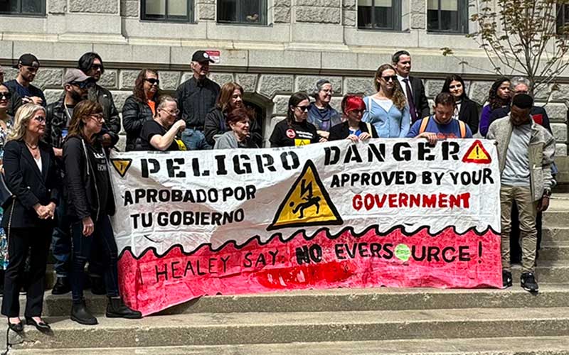 East Boston residents and advocates stand outsid court in a rally to stop the construction of a harmful electric substation. They carry a sign that reads peligro abrobado por tu gobierno in Spanish. On the right side text read in English: Danger approved by your government. At the bottom, a call to action to Massachussetts Governor. It reads: Healey say: No Eversource!