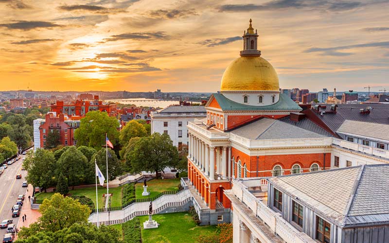 Massachusetts State House at sunset, featuring its iconic golden dome, surrounded by trees and city buildings, with a view of the Charles River in the background.