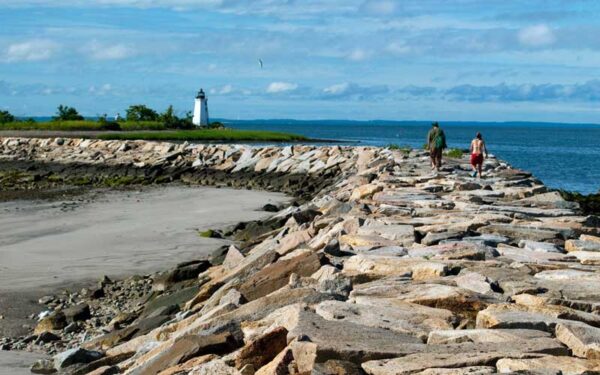 A rocky jetty stretches into the distance, guiding two people walking toward a quaint white lighthouse under a bright, sunny sky. The scene includes a sandy beach on one side and the blue waters of Long Island Sound on the other, evoking a serene coastal atmosphere.