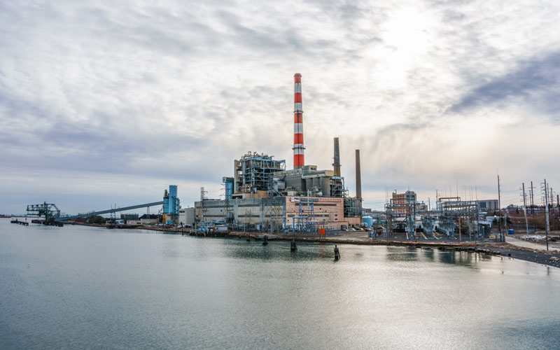 An industrial power plant with a tall red-and-white smokestack sits along the water in Bridgeport, Connecticut, under a cloudy sky. The facility’s industrial structures dominate the landscape, contrasting with the calm water and surrounding area.