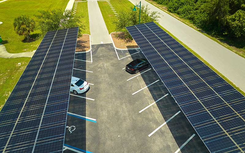 A parking lot featuring solar panel canopies providing shade for vehicles, with a white car and a black car parked underneath. The scene is surrounded by green grass, trees, and a road, showcasing how renewable energy solutions can improve communities and reduce pollution.