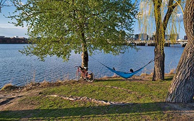 A person relaxes in a hammock by the Charles River with a bicycle parked nearby, showcasing the need for the Allston Multimodal Project to reconnect communities and provide better access to recreation and transportation.