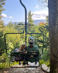 A mother and her child sit together on a ski lift, looking out at a scenic view of lush green trees and distant mountains.